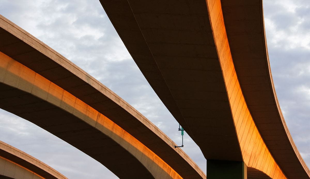 Aerial view of curved concrete highway overpasses bathed in warm golden light against a cloudy sky.