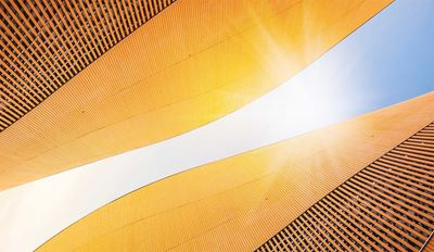 Upward-looking view of a modern building's curved golden-yellow exterior facade with horizontal ribbed panels, framed against a bright blue sky with sunlight