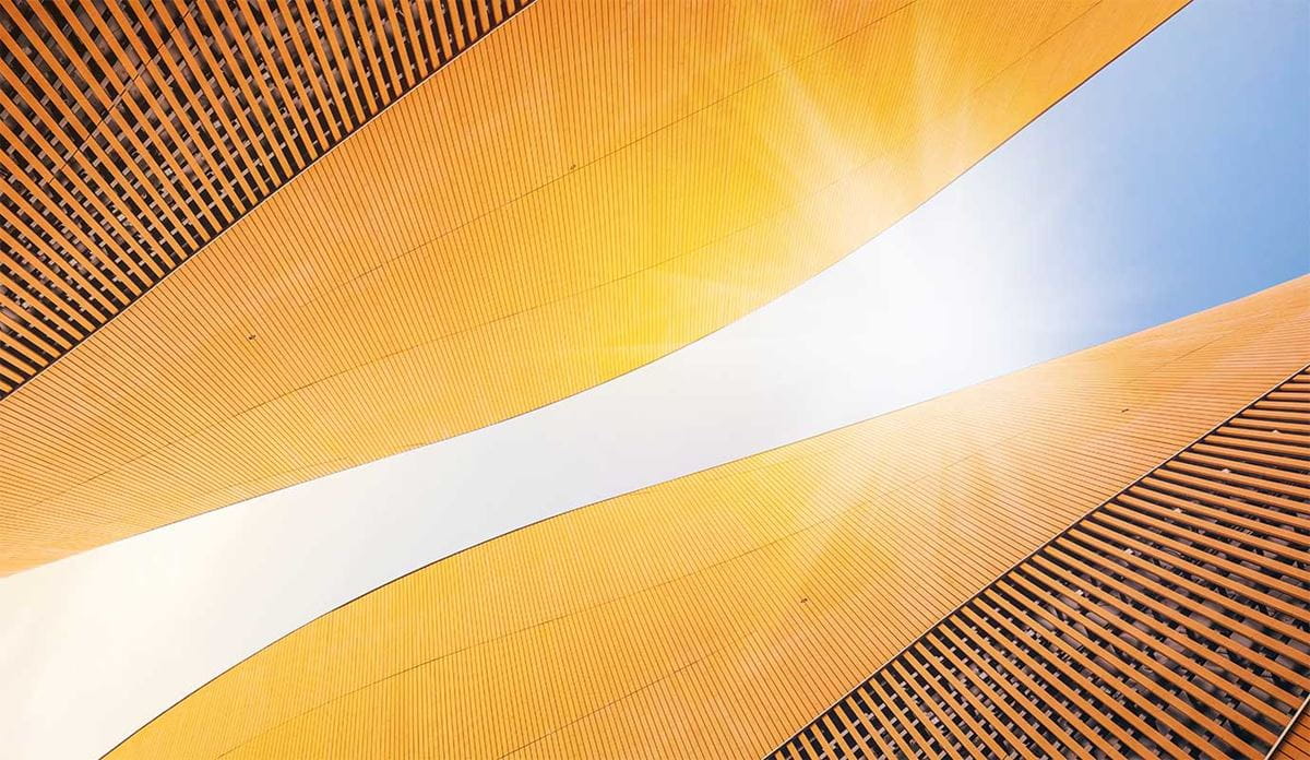 Upward-looking view of a modern building's curved golden-yellow exterior facade with horizontal ribbed panels, framed against a bright blue sky with sunlight